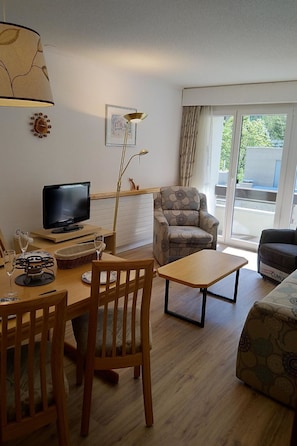 Ein Wohnzimmer mit Fernseher, Esstisch mit Stühlen, einer Couch, einer Lampe und einem Fenster mit Vorhängen.
