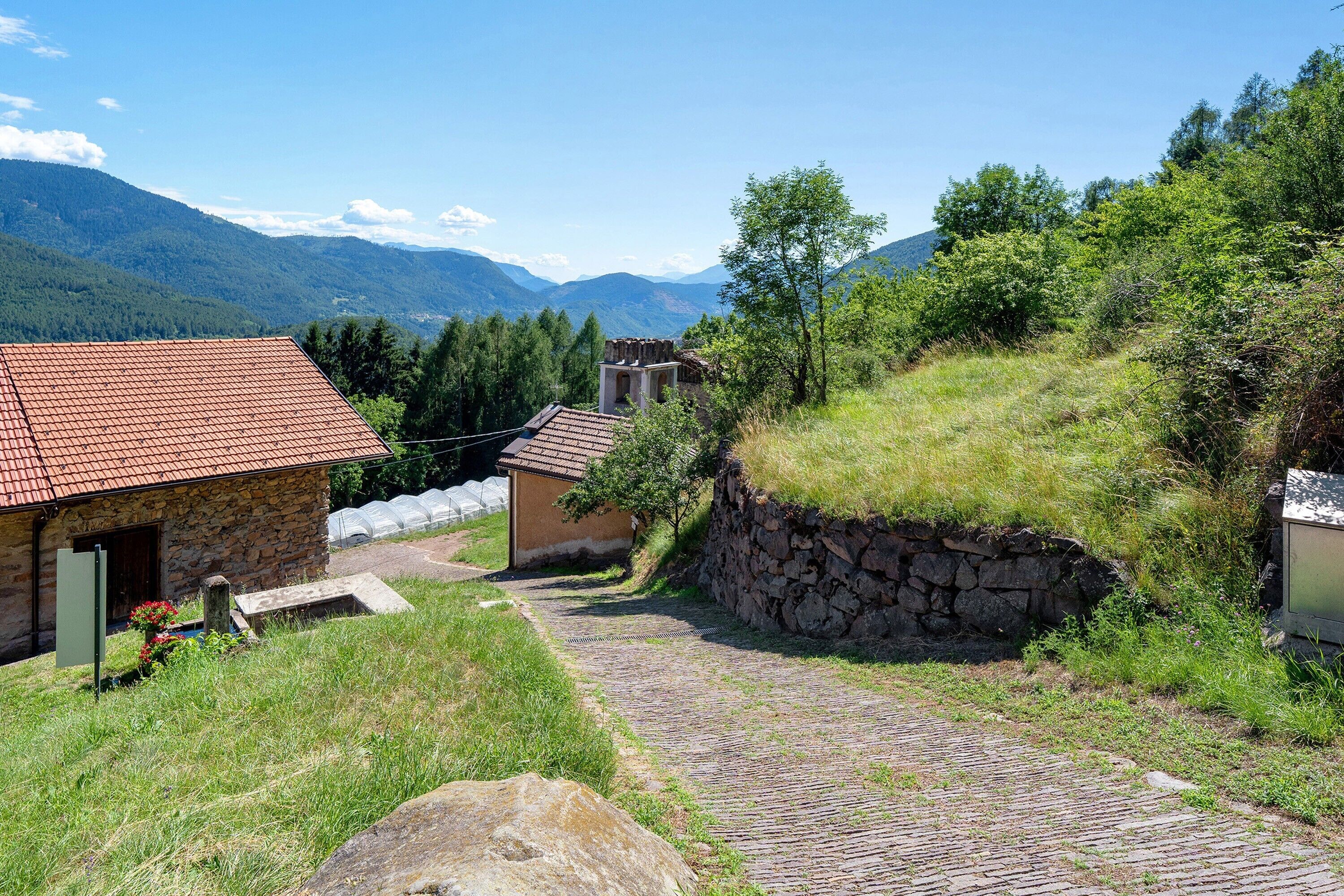 Un edificio de piedra con techo de tejas rojas, un camino adoquinado y montañas al fondo.