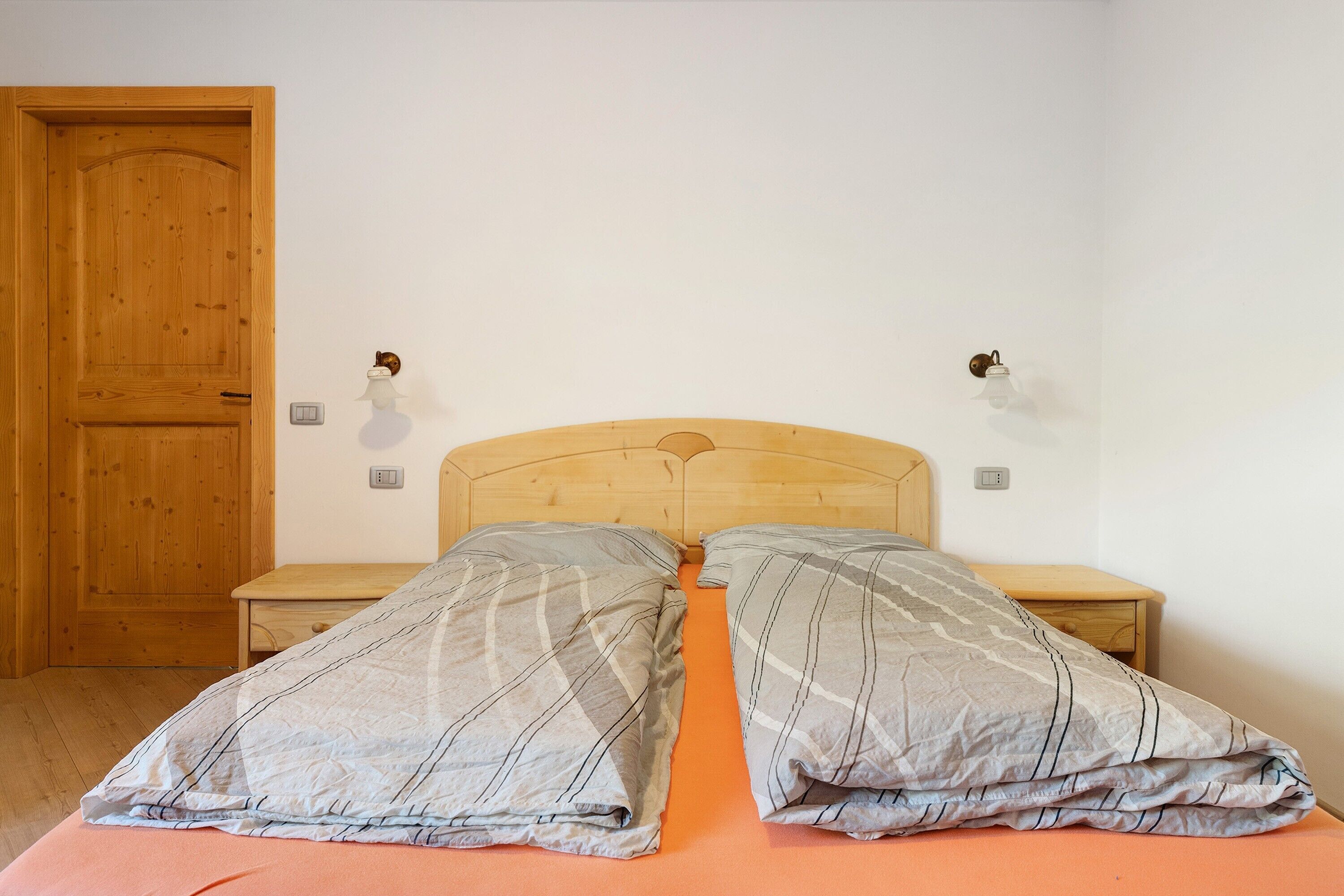 Ein Schlafzimmer mit Holztür, zwei Nachttischen, einem Bett mit grauen Streifen und einer schlichten weißen Wand.