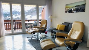Un salon moderne comprenant un canapé, deux fauteuils, une table basse en verre et une vue sur un paysage montagneux, visible à travers de grandes portes-fenêtres coulissantes.
