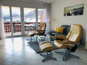 A modern living room with a sofa, two armchairs, a glass coffee table, and a view of a mountainous landscape through large sliding glass doors. - Edelweiss B 23 # 4-bed apartment, Dusche, WC 4-bed apartment, Dusche, WC by Interhome (Leukerbad)
