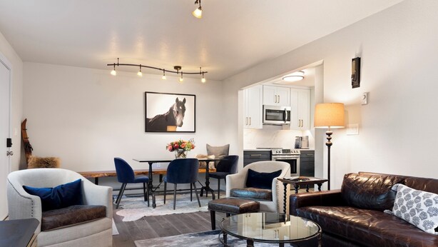 Una sala de estar moderna con un sofá, sillas, una mesa de centro de vidrio, un área de comedor con una mesa y sillas, y una cocina al fondo.