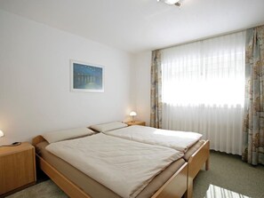 A hotel room with two beds, a nightstand, a lamp, a painting on the wall, and a window with curtains. - Salve 3 # 4-bed apartment, Dusche, WC 4-Bettwohnung, Dusche, WC by Interhome (Leukerbad)