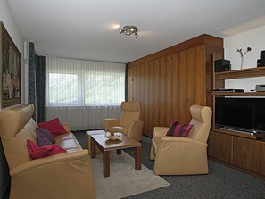 Ein möbliertes Wohnzimmer mit Sofa, Sesseln, Couchtisch, Fernseher und Holzschrank.