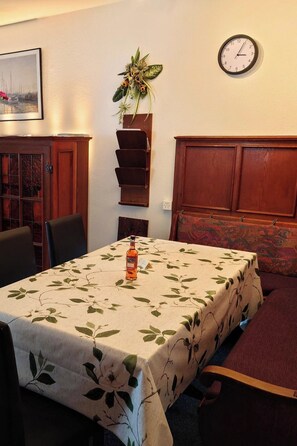 Ein Essbereich mit einem Tisch, der mit einer blumigen Tischdecke bedeckt ist, eine Spirituosenflasche, eine Wanduhr und ein Holzschrank.