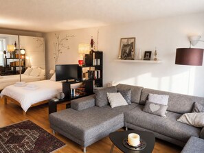A modern living room with a sofa, a television, a bookshelf, and a bed. - Majesta Leukerbad # 2-bed apartment, Dusche, WC by Interhome (Leukerbad)