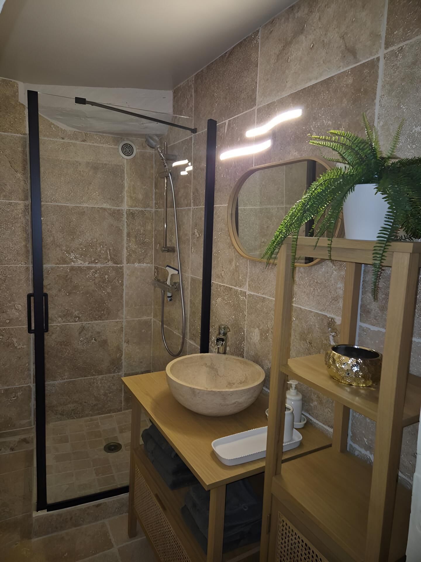 A bathroom with a glass-enclosed shower, a wooden vanity with a bowl sink, a mirror, and a shelf with a plant and decorative items.