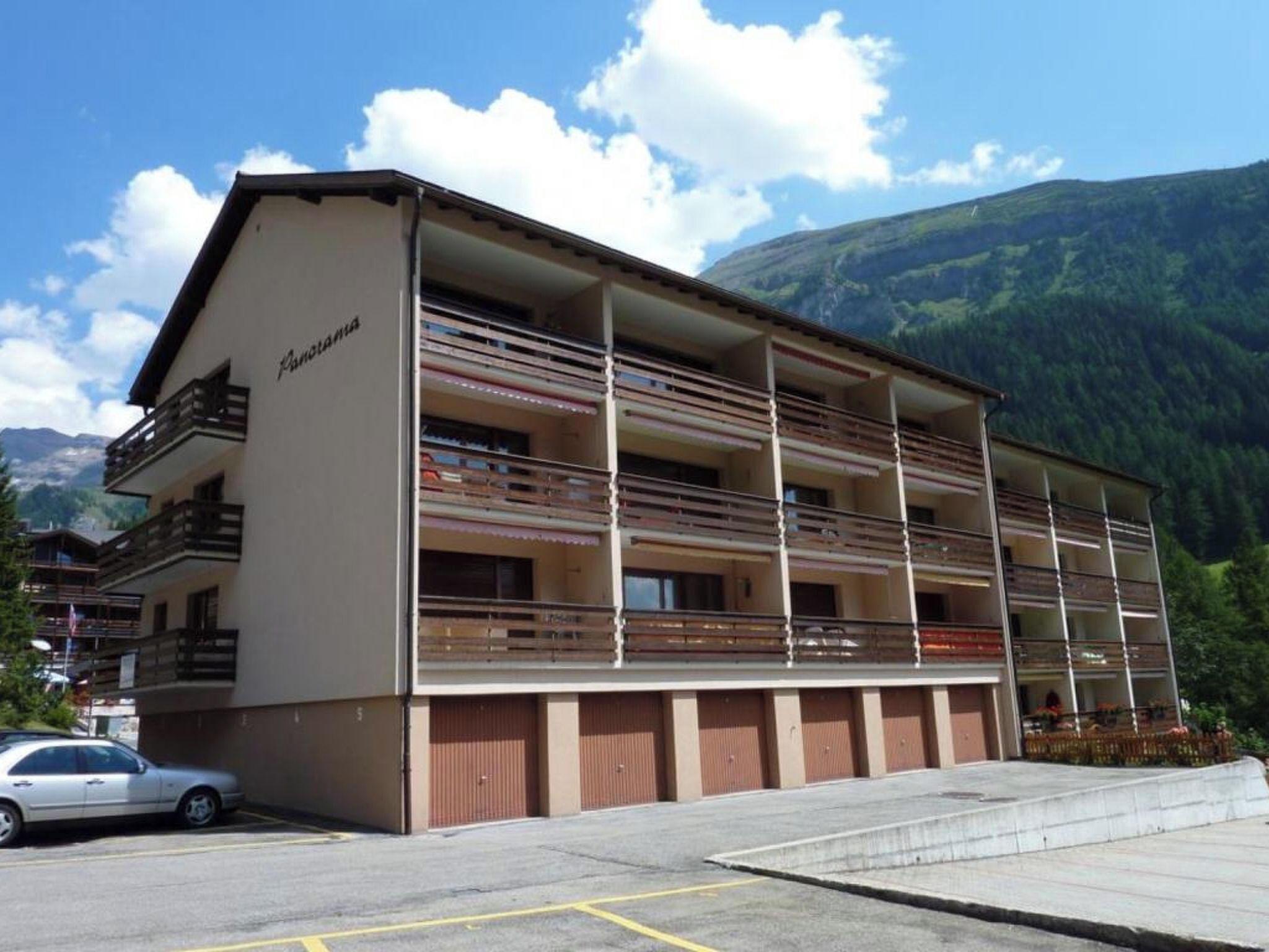 Un edificio de varios pisos con balcones, una zona de estacionamiento y montañas al fondo.