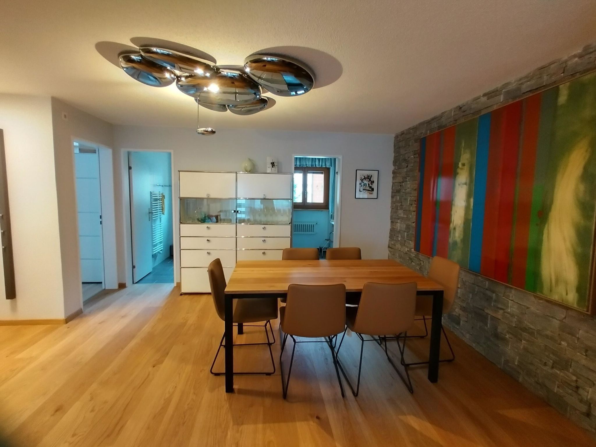 Una zona de menjador amb una taula i caires de fusta, un llum de sostre modern, una cuina al fons i una paret colorida.