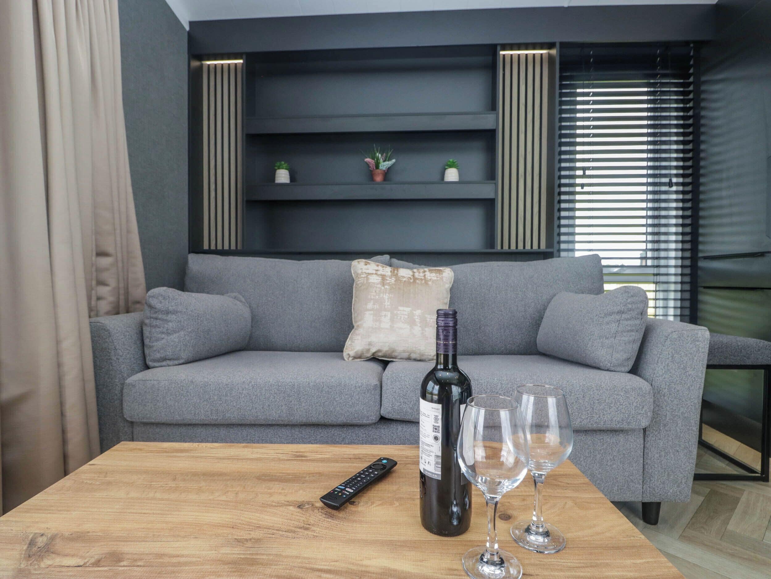 Una sala moderna con un sofá gris, una mesa de centro de madera, un control remoto, una botella de vino y dos copas de vino.