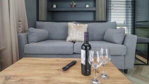 Una sala moderna con un sofá gris, una mesa de centro de madera, un control remoto, una botella de vino y dos copas de vino.