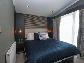 A bedroom with a bed, bedside tables, a radiator, and a window with curtains. - Heron (Millom)