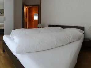 A neatly made bed in a hotel room with a wooden headboard, a bedside table with a lamp, and a door leading to another room. - Edelweiss E 13 # 4-bed apartment, Dusche, WC 4-bed apartment, Dusche, WC by Interhome (Leukerbad)