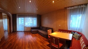 Ruime kamer met houten vloer, een bank, een eettafel met stoelen en grote ramen met gordijnen.