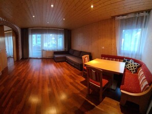 A spacious room with a wooden floor, a sofa, a dining table with chairs, and large windows with curtains. - Arca 2 # 5-bed apartment, Bad, WC 5-bed apartment, Bad, WC by Interhome (Leukerbad)