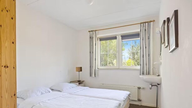 A bedroom with two beds, a window with curtains, a bedside lamp, a radiator, and a sink. - Romantic Villa for 2-guests With Sauna, Makkum (Makkum)