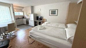 Habitació d'hotel petita amb un llit, un escriptori, una cadira, un televisor i una cuineta.