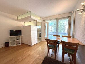 Un espace de vie compact comprenant un coin repas, un téléviseur et une cuisine visible à travers une porte ouverte.