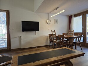A room with a wooden dining table, chairs, a mounted TV, a radiator, and a clock on the wall. - Fortuna 316 # 5-bed apartment, Dusche, WC 5-Bettwohnung, Dusche, WC by Interhome (Leukerbad)