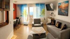 Ein modernes Wohnzimmer mit einem grauen Sofa, einem kleinen Tisch, einem an der Wand befestigten Fernseher und Blick ins Freie durch ein großes Fenster.