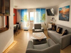 Ein modernes Wohnzimmer mit einem grauen Sofa, einem kleinen Tisch, einem an der Wand befestigten Fernseher und Blick ins Freie durch ein großes Fenster.