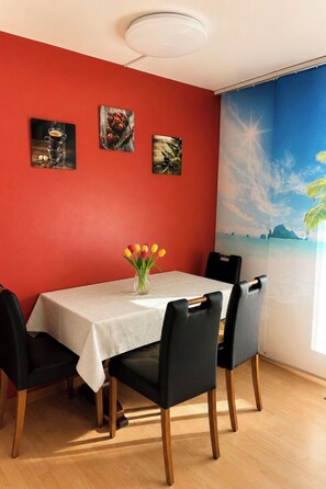 Ein Essbereich mit roter Wand, einem Tisch mit weißer Tischdecke, vier Stühlen und einer Vase mit Tulpen.