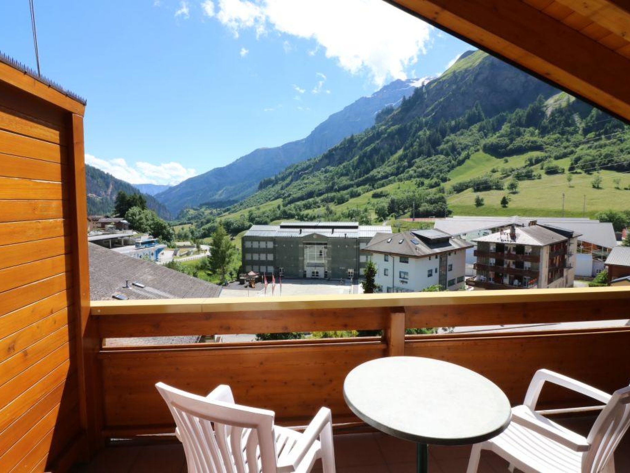 A wooden balcony with a table and chairs, overlooking a mountainous landscape with buildings.