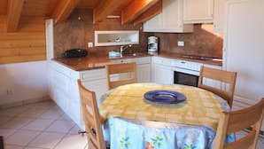 Een compacte keuken met houten balken, witte kastjes, een eettafel met een bloemrijk tafelkleed en een koffiezetapparaat.