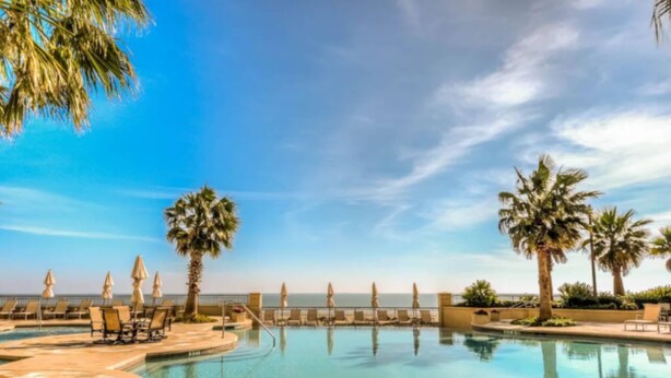 A pool with lounge chairs and palm trees, surrounded by a clear sky. - Palisade Palms Beach High Rise (Galveston)