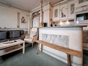 Een woonkamer met een bank, een houten zitbankje, een open haard en een keuken met een fornuis.