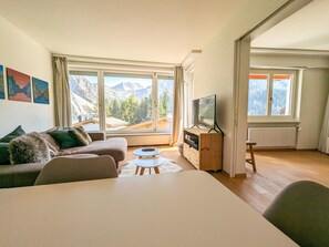 Un salon moderne doté d’une grande fenêtre donnant sur les montagnes, d’un canapé, d’un téléviseur et d’une petite table.