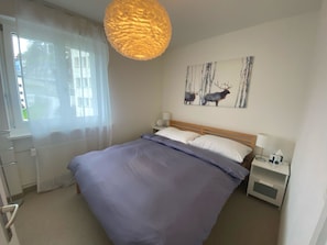 Une chambre à coucher avec un grand lit, une table de chevet avec une lampe, une fenêtre ornée de rideaux légers et une œuvre d’art représentant un élan, accrochée au mur.