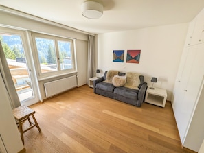 A spacious living room with a grey sofa, two side tables, a radiator, and a window with a view of trees. - Viktoria A39 by Arosa Holiday Appartement/Fewo, Dusche, WC, 6-Bettwohnung by Interhome (Arosa)