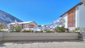Une terrasse avec un sol carrelé, des montagnes enneigées en arrière-plan et des bâtiments dotés de balcons.