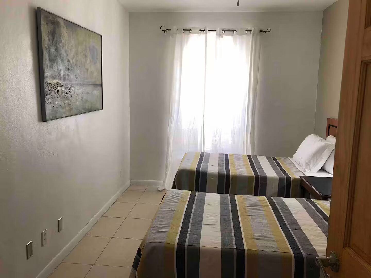 A bedroom with two beds, a window with curtains, a painting on the wall, and a door.