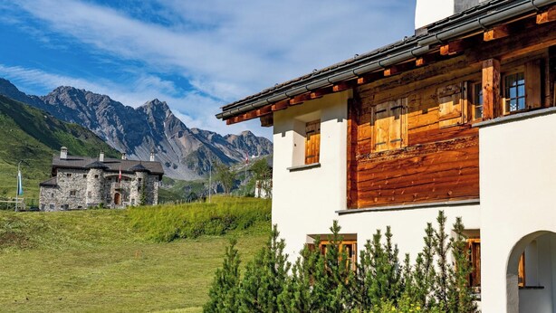 Un edificio de dos pisos con contraventanas de madera y fachada de piedra, con montañas al fondo.