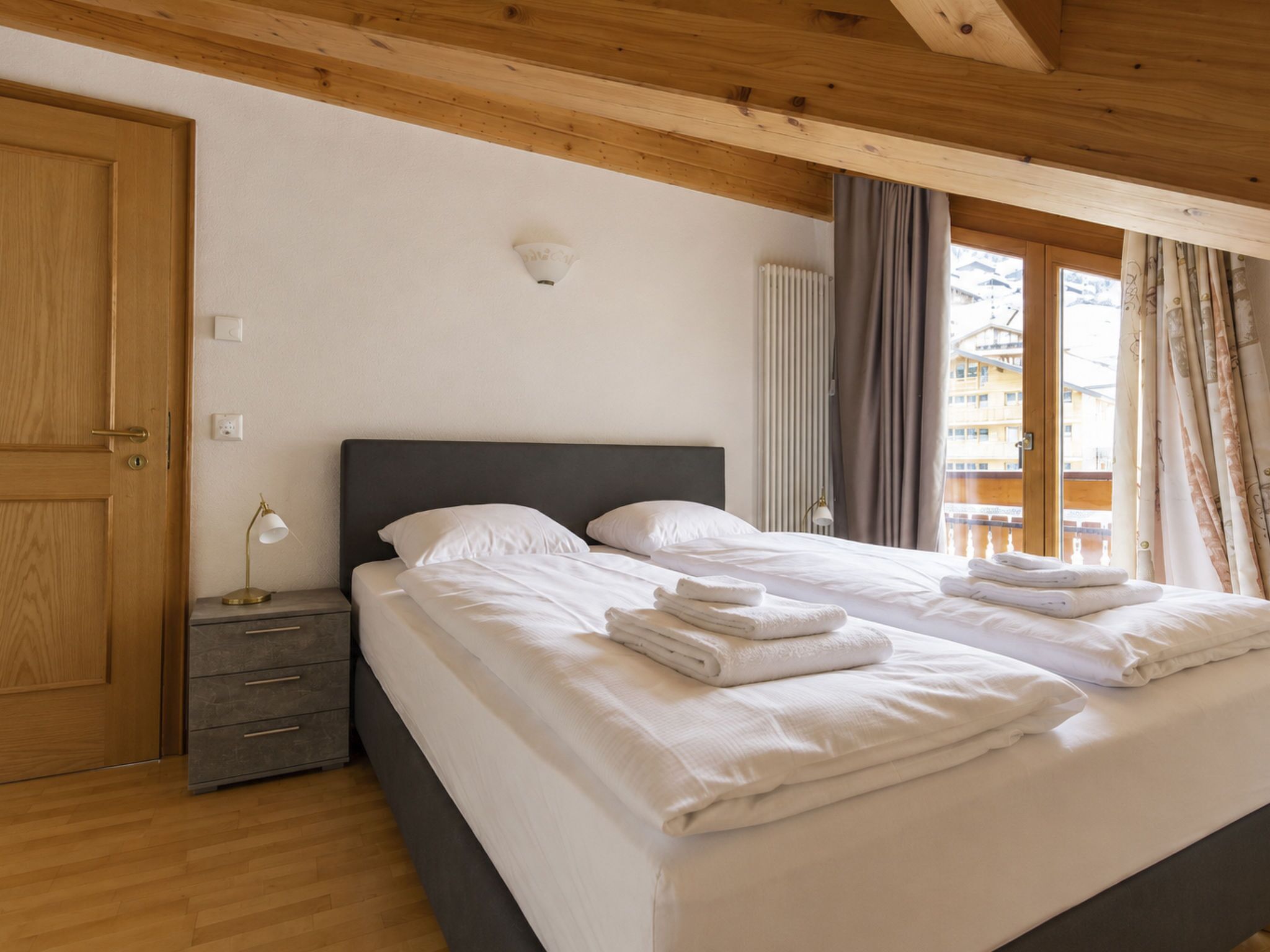 Une chambre à coucher avec un lit double, des tables de chevet, une lampe, un radiateur et une vue sur un immeuble par la fenêtre.