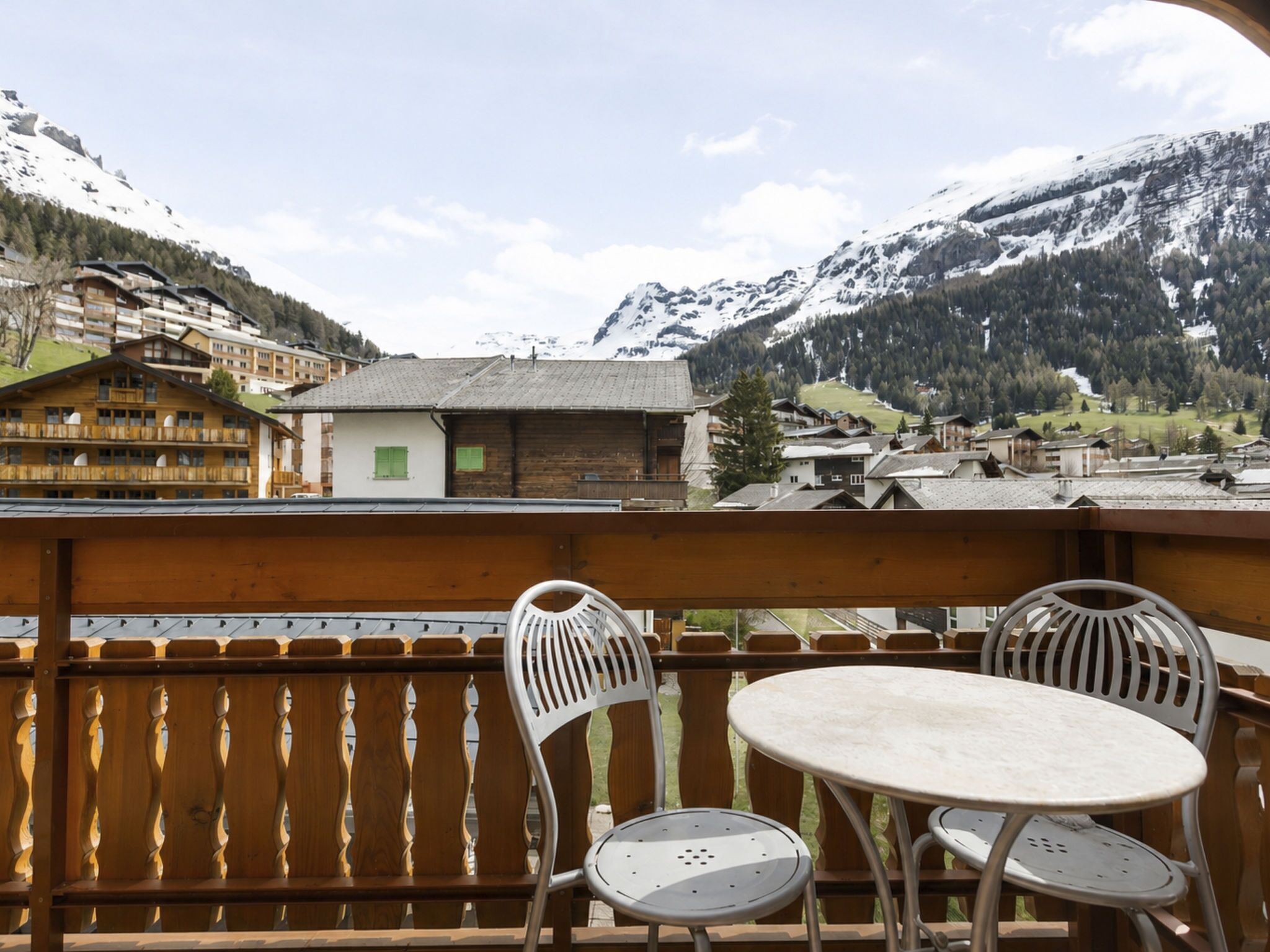 A balcony with two chairs and a table, overlooking a mountainous landscape with buildings.