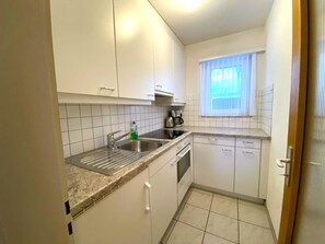 Eine kompakte Küche mit weißen Schränken, einer Edelstahl-Spüle und einem Fenster mit Jalousien.
