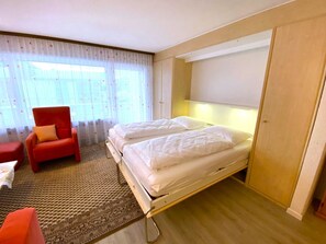Une chambre d’hôtel avec un lit, une chaise rouge, une petite table et une grande fenêtre avec des rideaux.