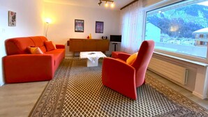 Ein Wohnzimmer mit einer Couch, einem Sessel und einem Couchtisch. Im Raum befindet sich ein Fernseher und ein Schrank.