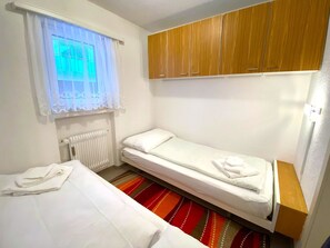Ein kleines Zimmer mit einem Einzelbett, einem Fenster, einer Heizung, einem Holzschrank und einem Streifenmuster-Teppich.