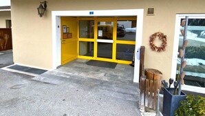 Ein Gebäudeeingang mit gelber Tür, einem Schild mit der Zimmernummer 2 und einem Kranz auf der rechten Seite.