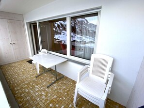 Ein kleiner Raum mit einem weißen Tisch und zwei weißen Stühlen, ein Fenster mit Blick auf schneebedeckte Gebäude und ein Fliesenboden.