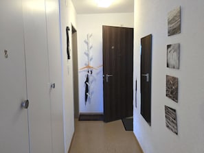 A hallway with a white door, a wooden door, and a coat rack with a coat and umbrella. - Leuca 24 # 2-bed apartment, Dusche, WC 2-Bettwohnung, Dusche, WC by Interhome (Leukerbad)