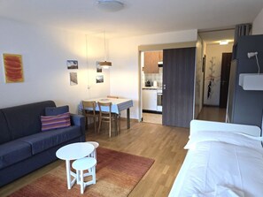 Un espacio habitable compacto con sofá, una pequeña mesa de comedor, una cama y una kitchenette al fondo.