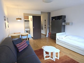 A modern living room with a sofa, a small dining table, a white bed, and a kitchen area in the background. - Leuca 24 # 2-bed apartment, Dusche, WC 2-Bettwohnung, Dusche, WC by Interhome (Leukerbad)