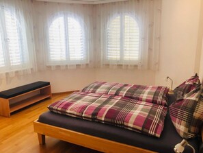 A bedroom with a bed, a bench, and three windows with curtains. - Edelweiss C 11 # 4-bed apartment, Dusche und Bad, WC by Interhome (Leukerbad)