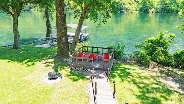 Um deck à beira do lago com cadeiras vermelhas, um pequeno píer e uma área gramada com árvores.