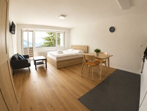 Ein Schlafzimmer mit Bett, Schreibtisch, Stuhl, Fernseher und Blick auf Berge durch ein Fenster.